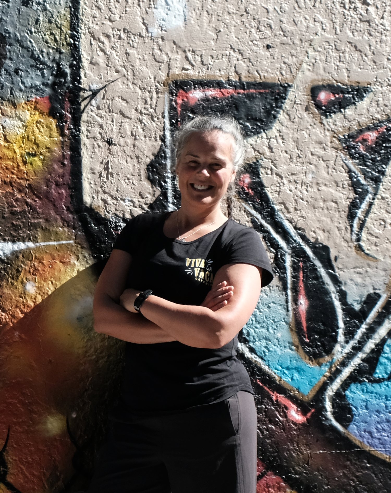 A woman smiling with crossed arms standing in front of colorful graffiti on a textured wall. She is the owner of the business, event caterer Chelsea.