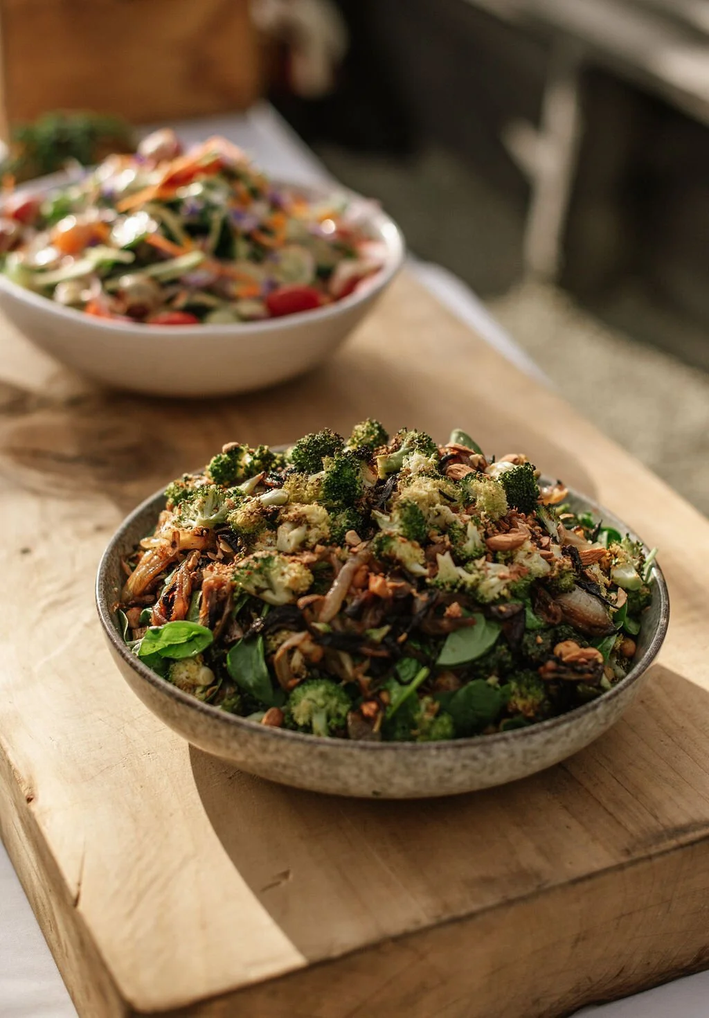 A bowl of salad with broccoli, spinach, shredded chicken, and toasted nuts on a wooden board, with another bowl of salad in the background, showcasing catering options.
