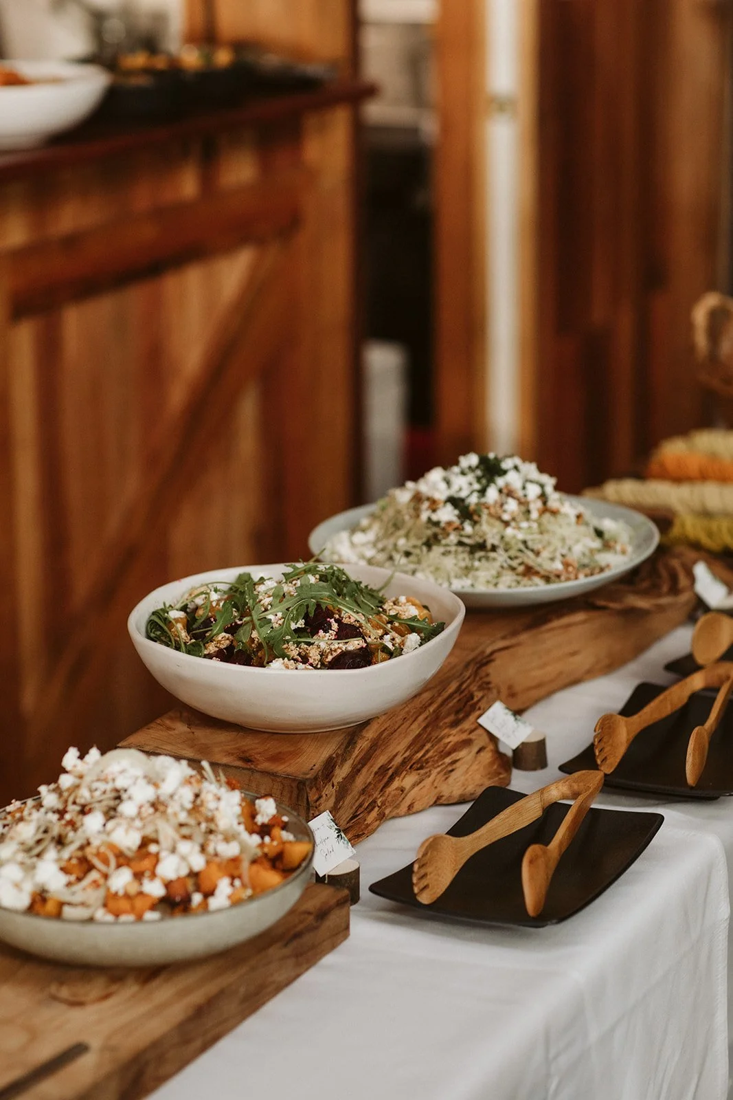 Bowls of salad on a wooden serving board with black empty plates and wooden utensils on a white tablecloth.