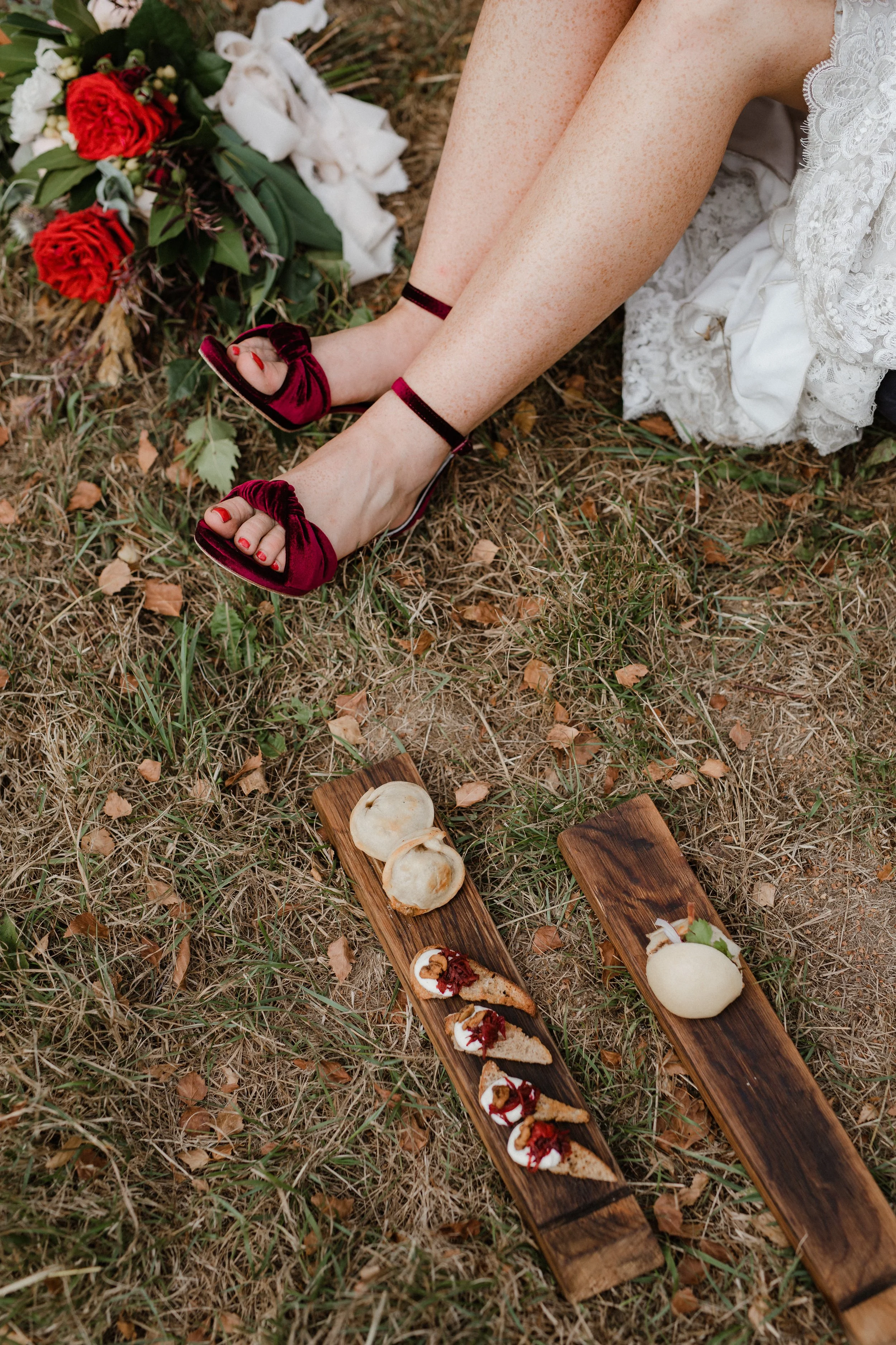 Person in a white lace dress sitting on grassy ground with red velvet shoes, feet crossed, and a bouquet of red and white flowers nearby. Two wooden boards with various desserts, including meringues and cake slices, are on the ground.
