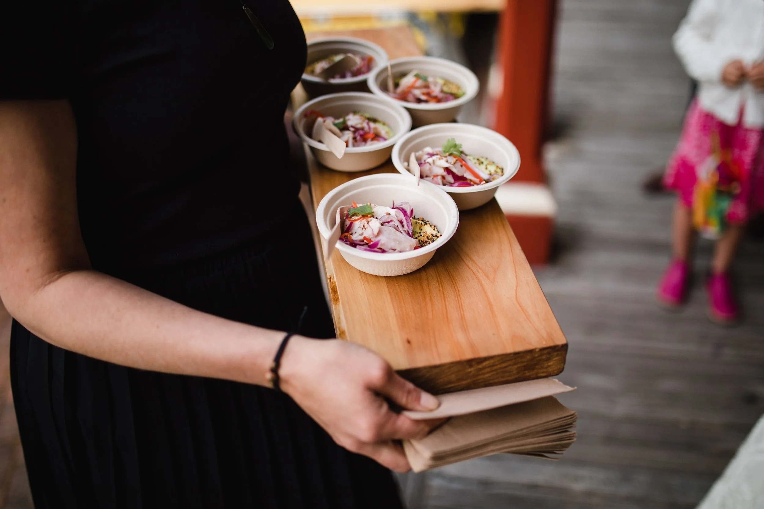 A person holding a wooden tray with six small bowls of food, likely at an event where food has been catered, with a child in colorful clothing in the background.