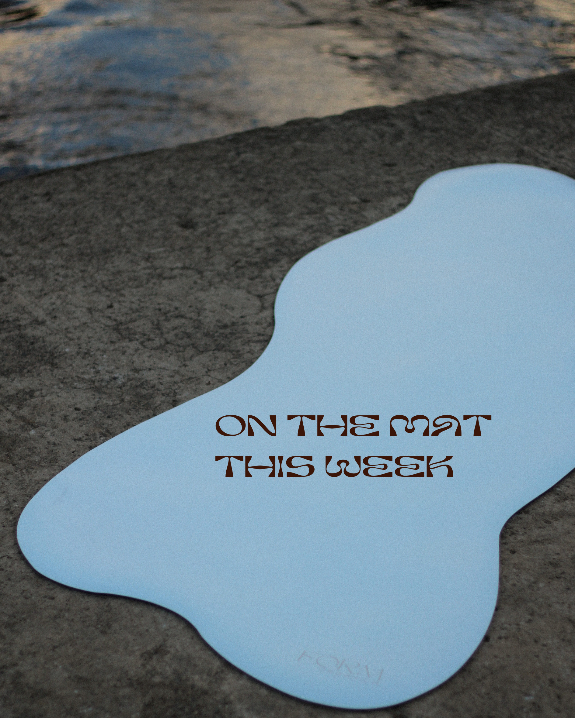 A puddle of water on the ground with a reflection, with a paper note on the surface reading 'On the mat this week'.