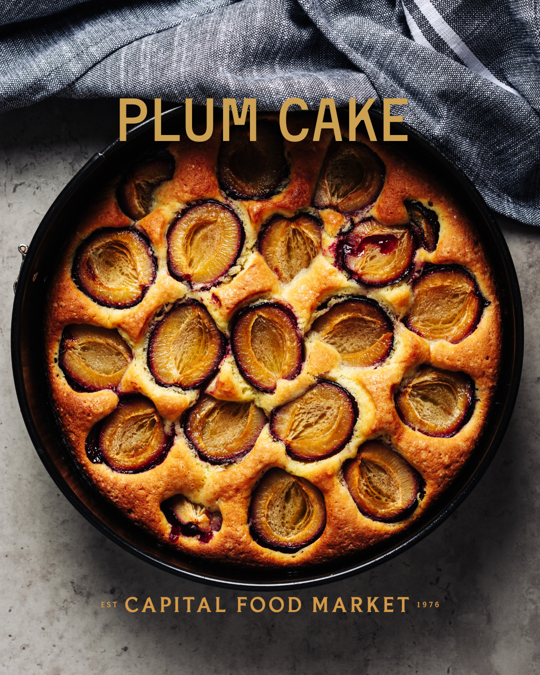 A round plum cake with halved plums on top, baked to a golden brown, in a black baking dish with a gray cloth partially covering the top edge.