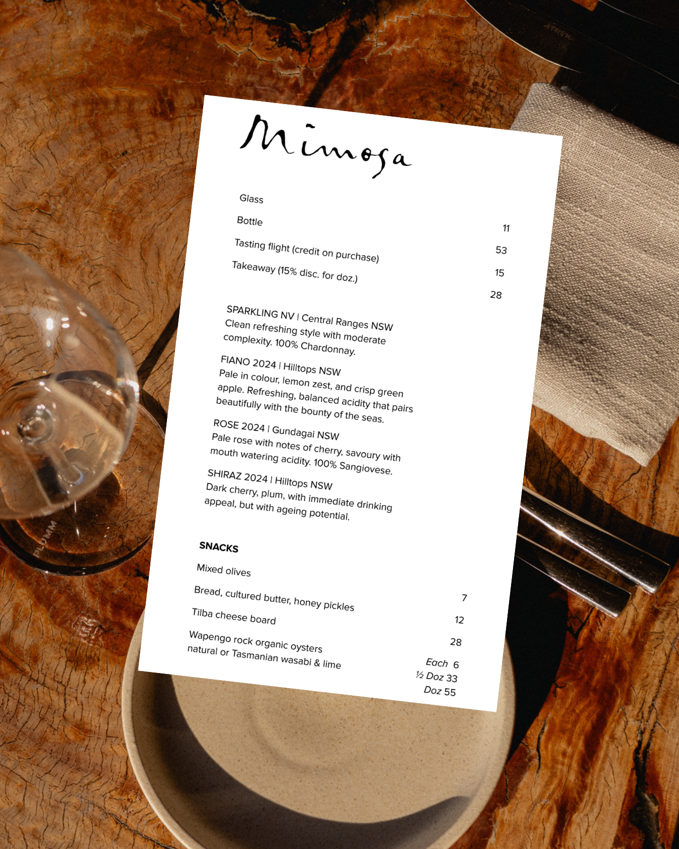 A menu titled 'Minoga' placed on a rustic wooden table, accompanied by a glass of white wine and an empty beige ceramic bowl with a fork and knife resting on a linen napkin.