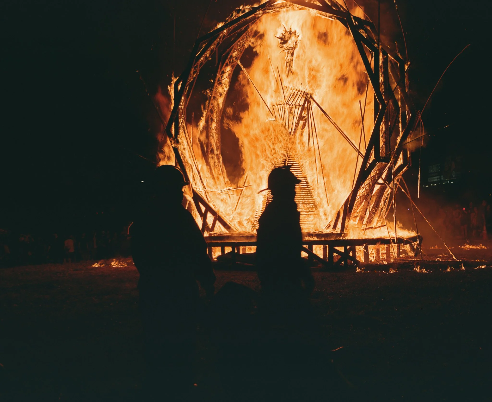 A large structure engulfed in flames, with silhouetted people standing nearby during a fire.