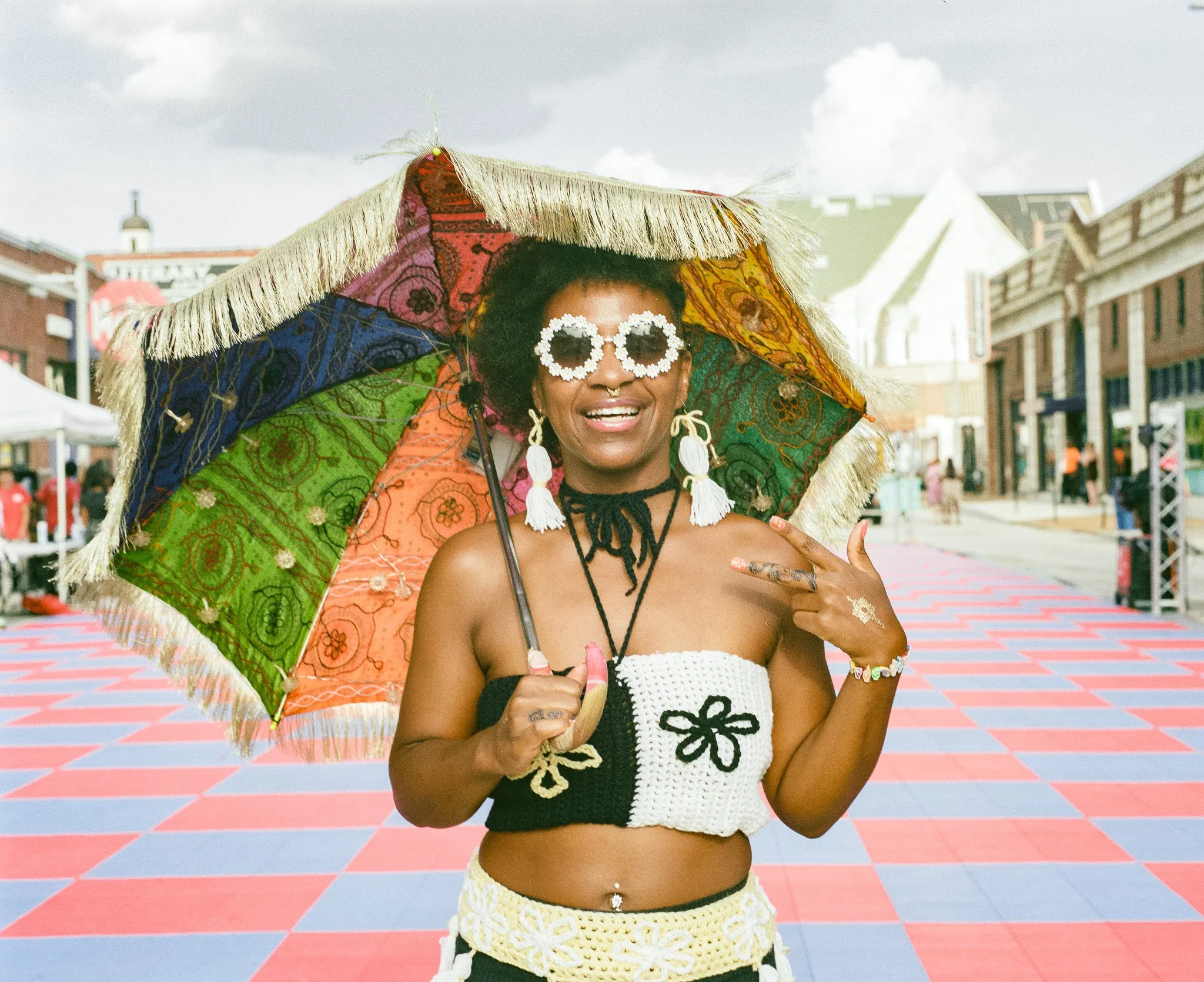 A person smiling, holding a colorful umbrella with rainbow and paisley patterns, standing on a patterned outdoor floor with buildings and a cloudy sky in the background.