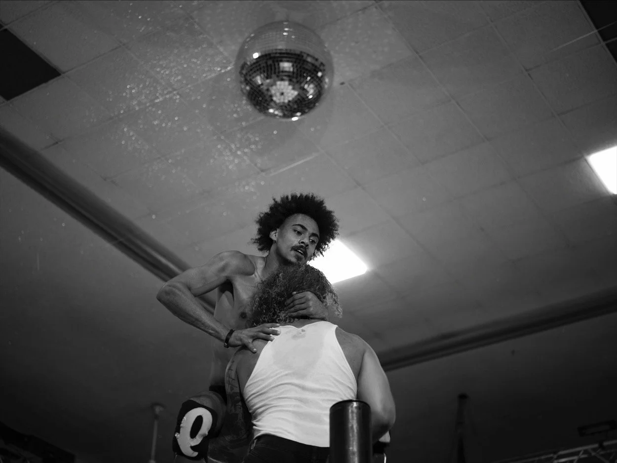 Black and white photo of a shirtless man with curly hair holding a woman from behind, both in a nightclub or dance environment. Disco ball overhead.