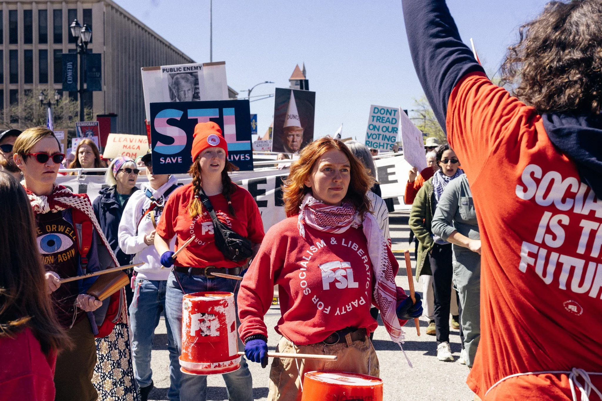 Protest in a city street with people holding signs and drums, some signs have political messages and images of Donald Trump, with buildings and streetlights in the background.
