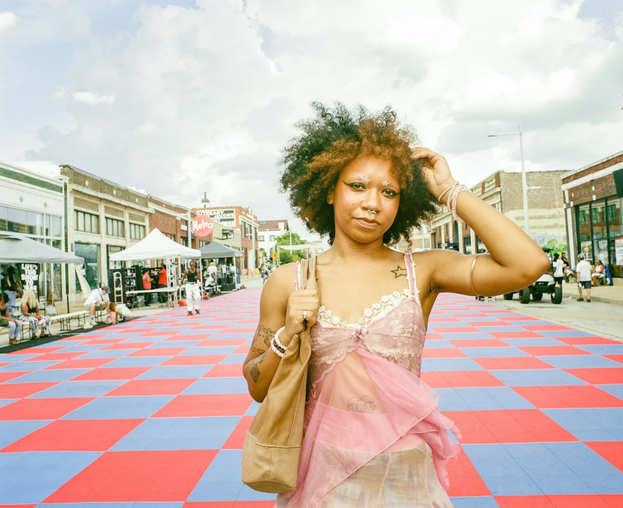 A young woman with curly hair, wearing a pink lace dress and carrying a tan bag, stands on a vibrant patterned outdoor area with tents and shops in the background, during daytime.
