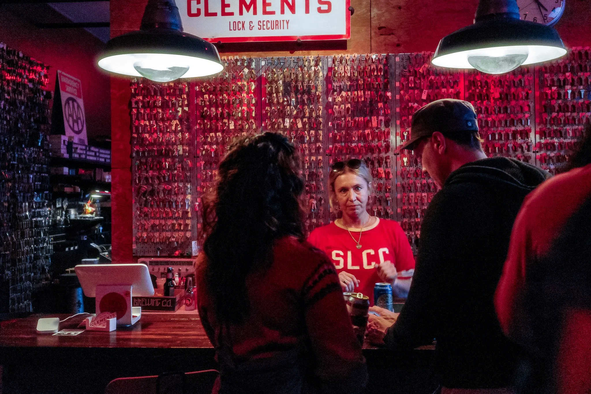 Several people are gathered in a dimly lit room with red lighting, engaging in conversation. The background features a wall filled with numerous keys hanging on hooks behind a woman wearing a red shirt. There are two large black pendant lights hangin