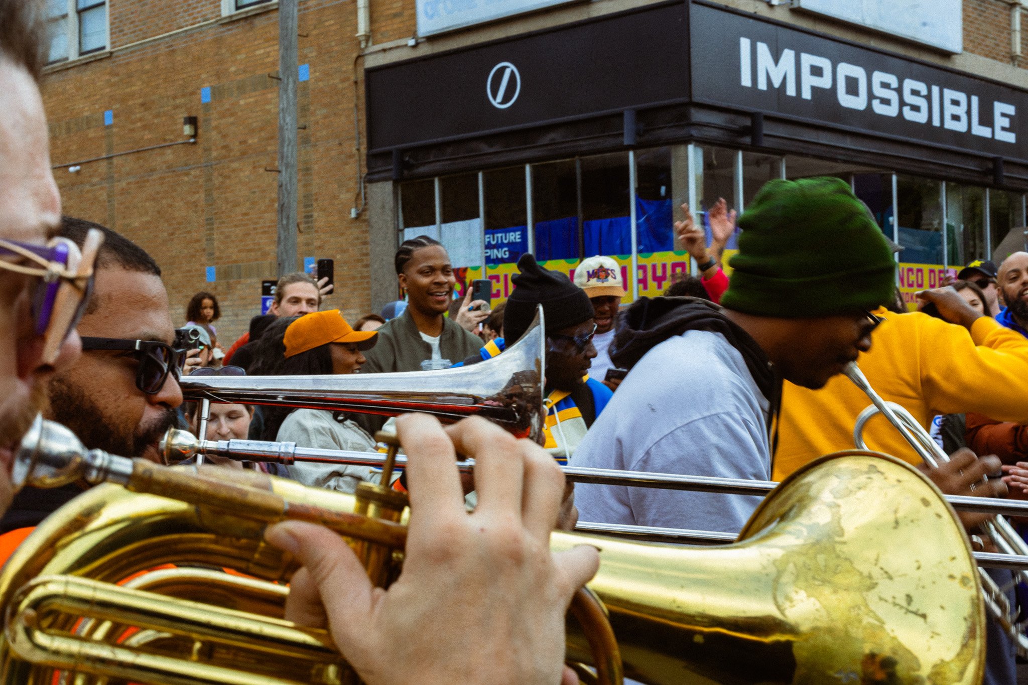 A street scene with a crowd of people, some taking photos, and a musicians playing brass instruments like trombones. The setting appears to be an outdoor event or parade with a building in the background showing a sign that reads 'IMPOSSIBLE'.
