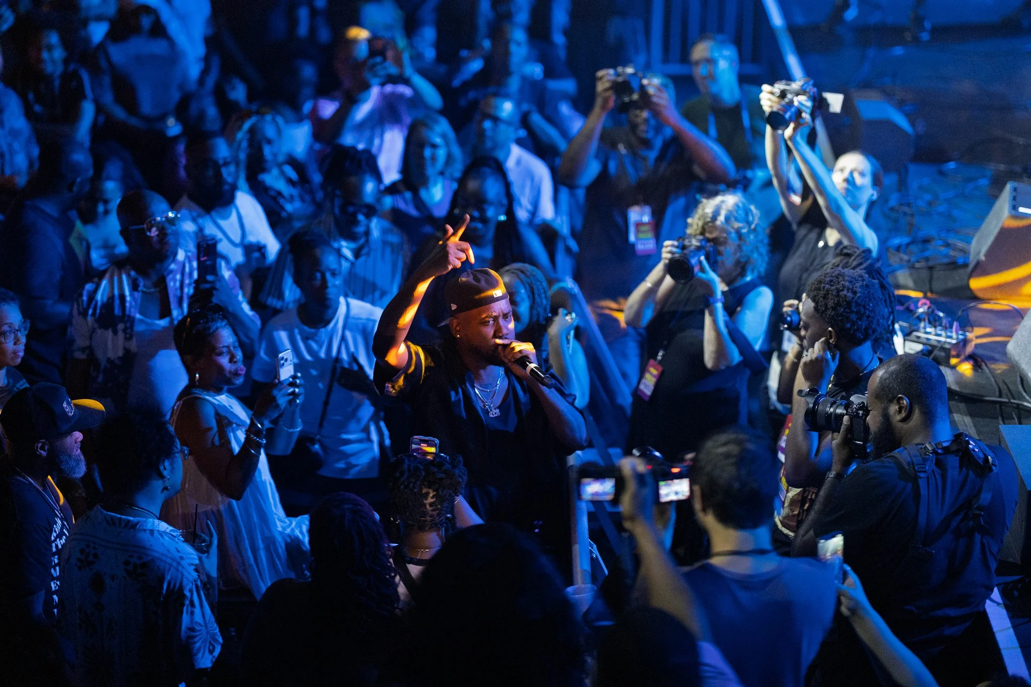 A group of people gathered together in a dark room, some are taking photographs or videos, and a woman in the foreground is speaking into a microphone with her hand raised. The lighting is blue.