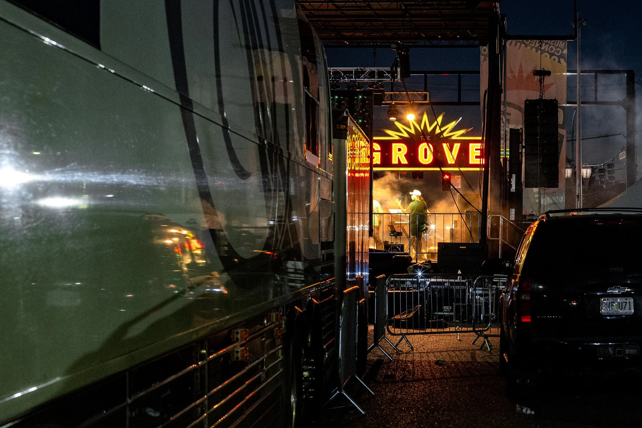 Nighttime scene at a music festival with a stage, bright neon sign reading 'GROVE,' and people performing on stage surrounded by loudspeakers and equipment, with parked cars and containers in the foreground.