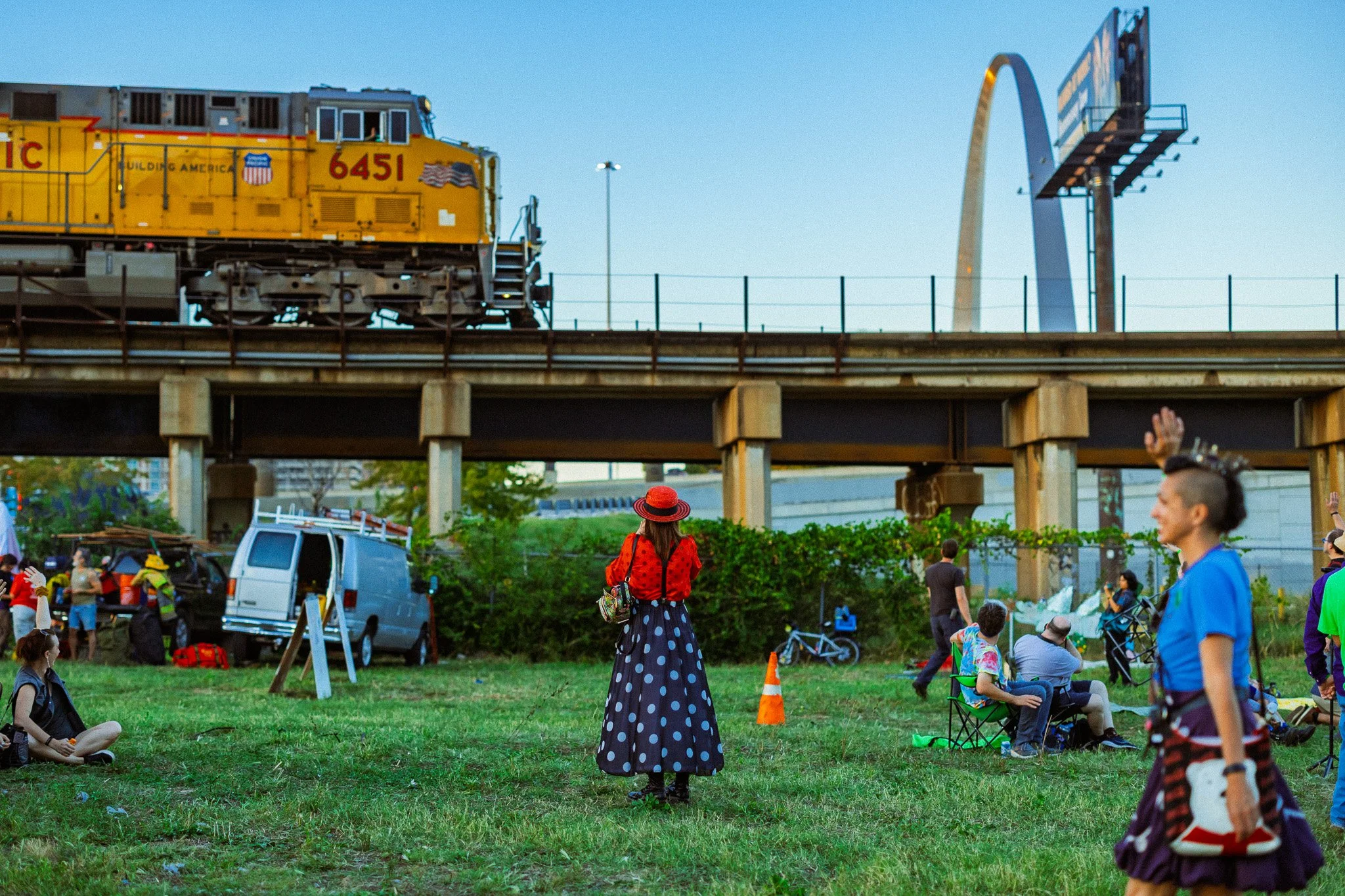 People gather outdoors in a grassy area under a bridge with a train passing overhead, some sitting on the ground, others standing or walking, with a woman in a polka dot skirt and hat in the center facing the train.