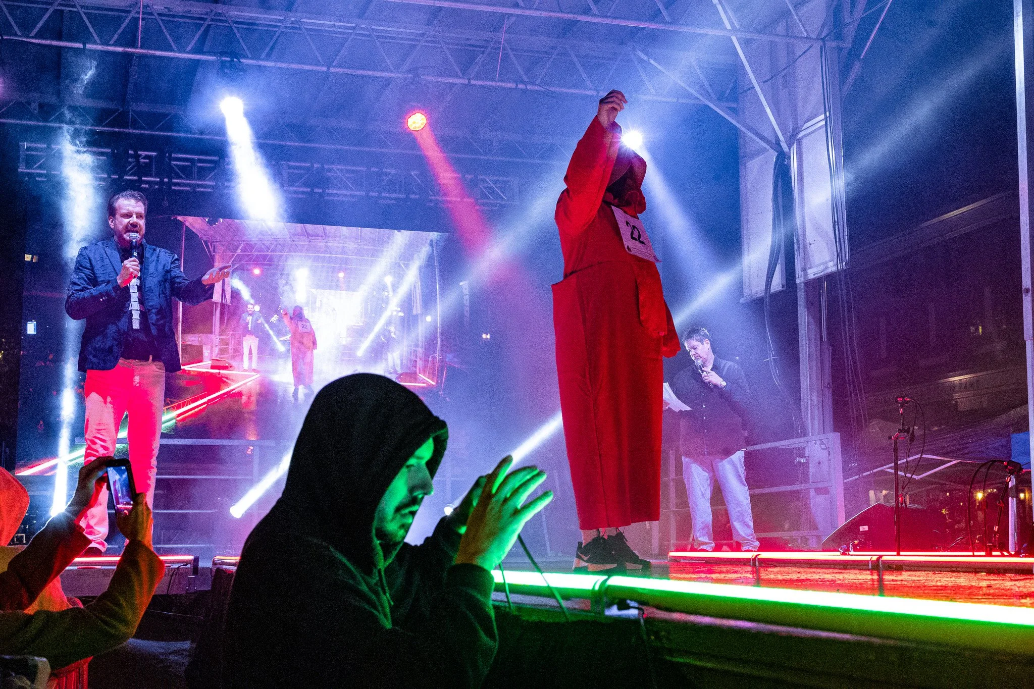 A stage with bright colorful lights, a man in a black jacket and pink pants holding a microphone, a person in a red outfit with a hood and number 22 on their chest standing with their face covered, and other performers in the background. Audience mem