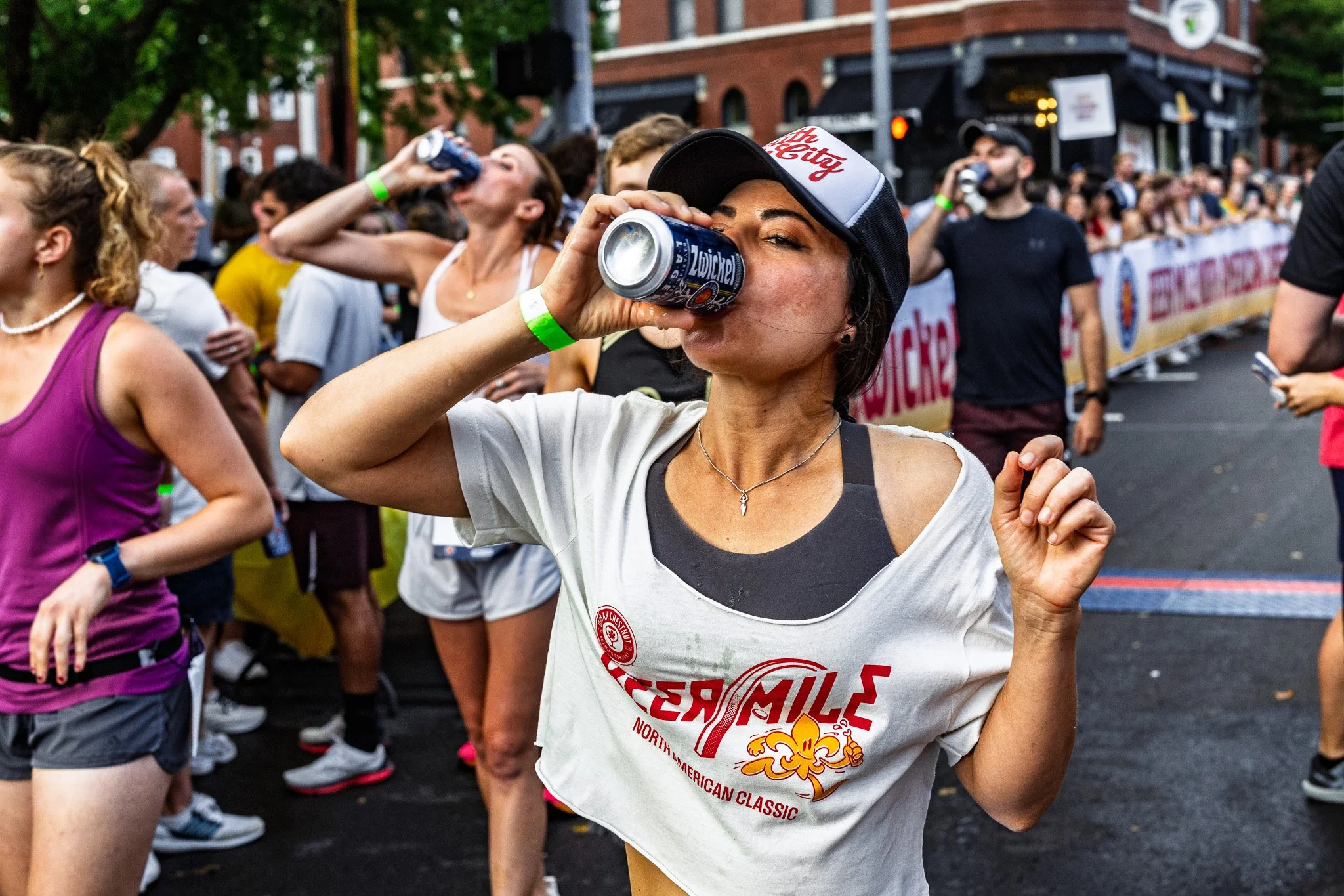 A group of people celebrating at an outdoor event, with a woman in the foreground wearing a white T-shirt and a cap, drinking from a soda can.