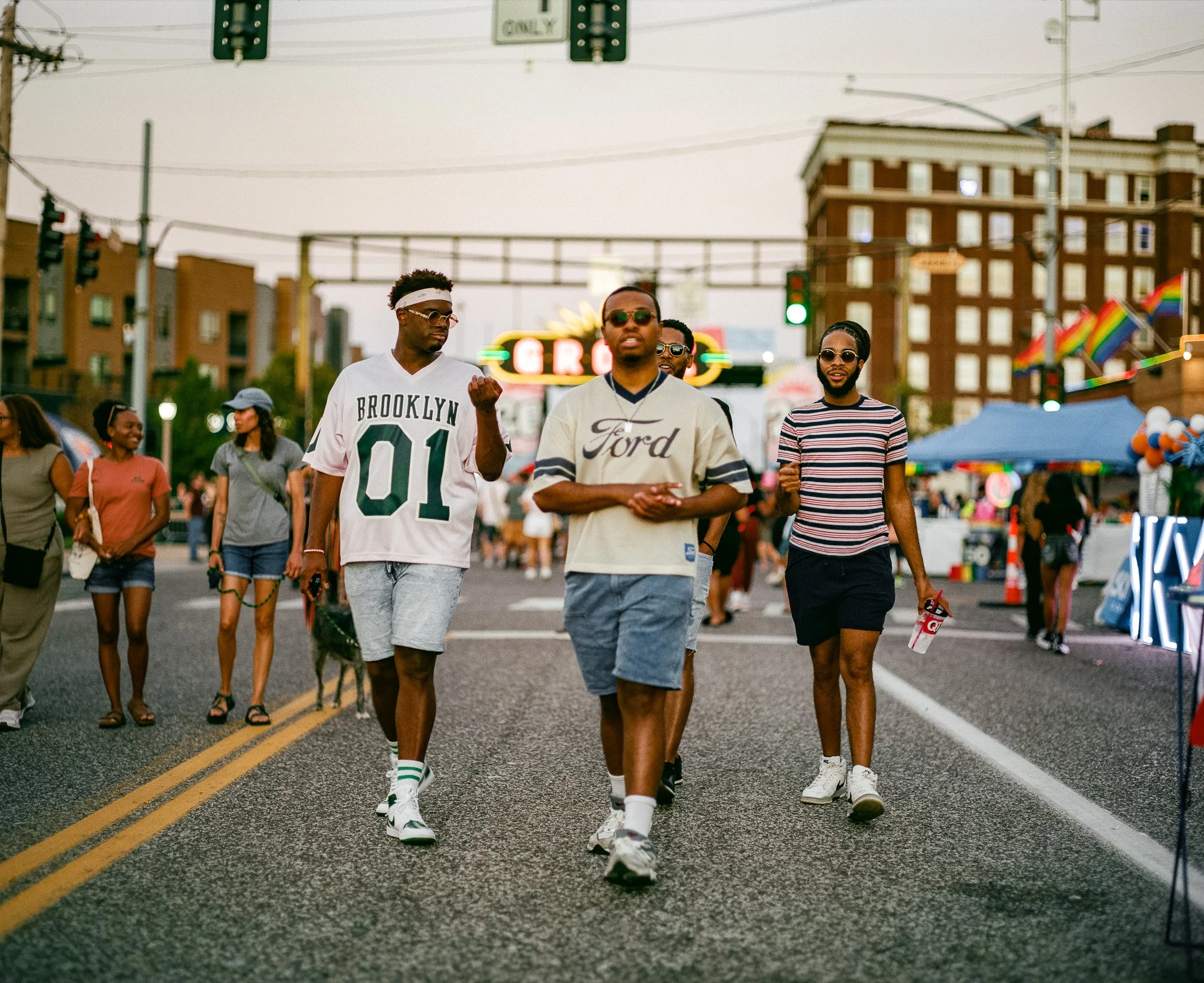 A group of young men walking on a street fair or festival, with booths, flags, and colorful lights around them. One man is wearing a Brooklyn sports jersey, and another is wearing a Ford sweatshirt, both wearing sunglasses.