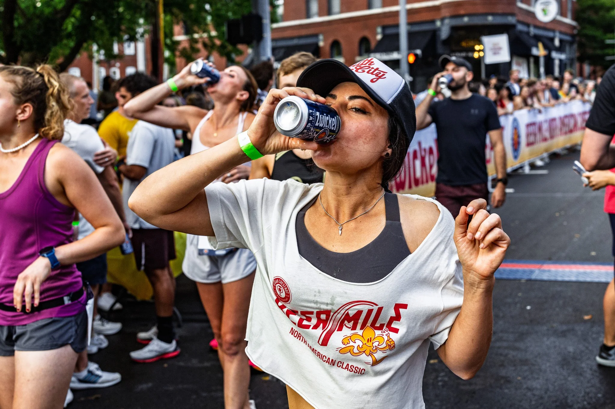 Group of people at an outdoor event, some drinking from cans, with a woman in the foreground wearing a white T-shirt and a black cap, taking a sip from a can.