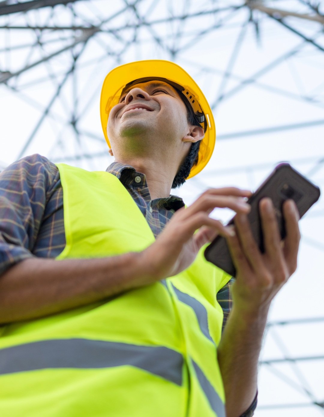A construction worker wearing a yellow hard hat and high-visibility vest, smiling and looking up at a metal structure, while holding a tablet.