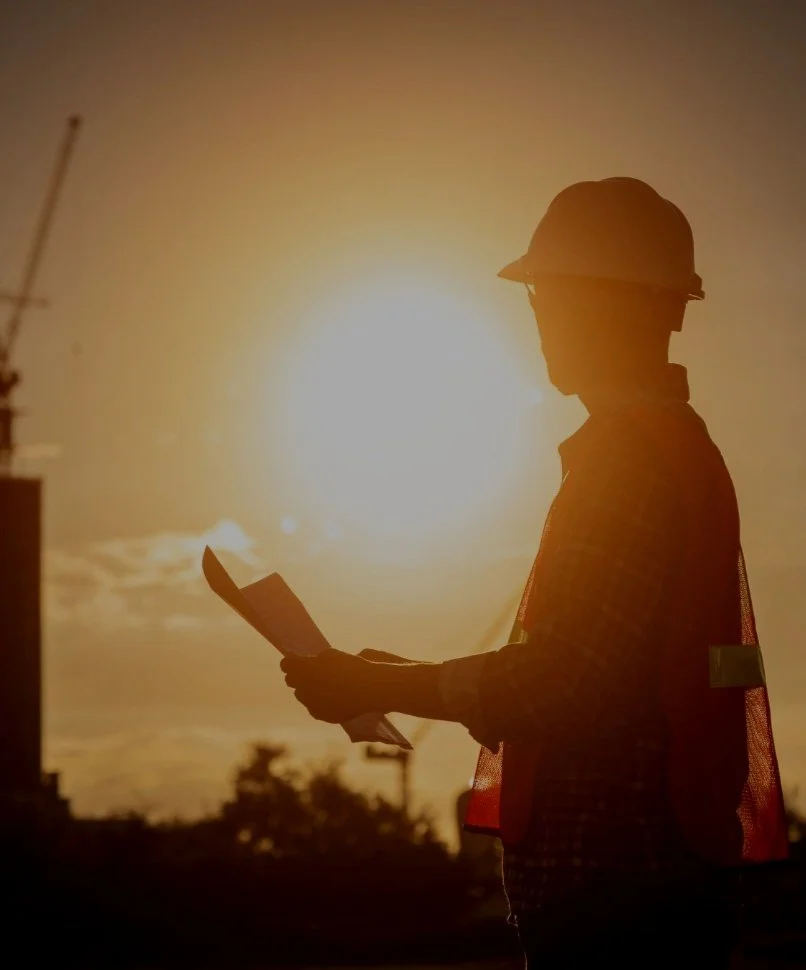 Silhouette of a construction worker wearing a hard hat and safety vest, reading a document during sunset.