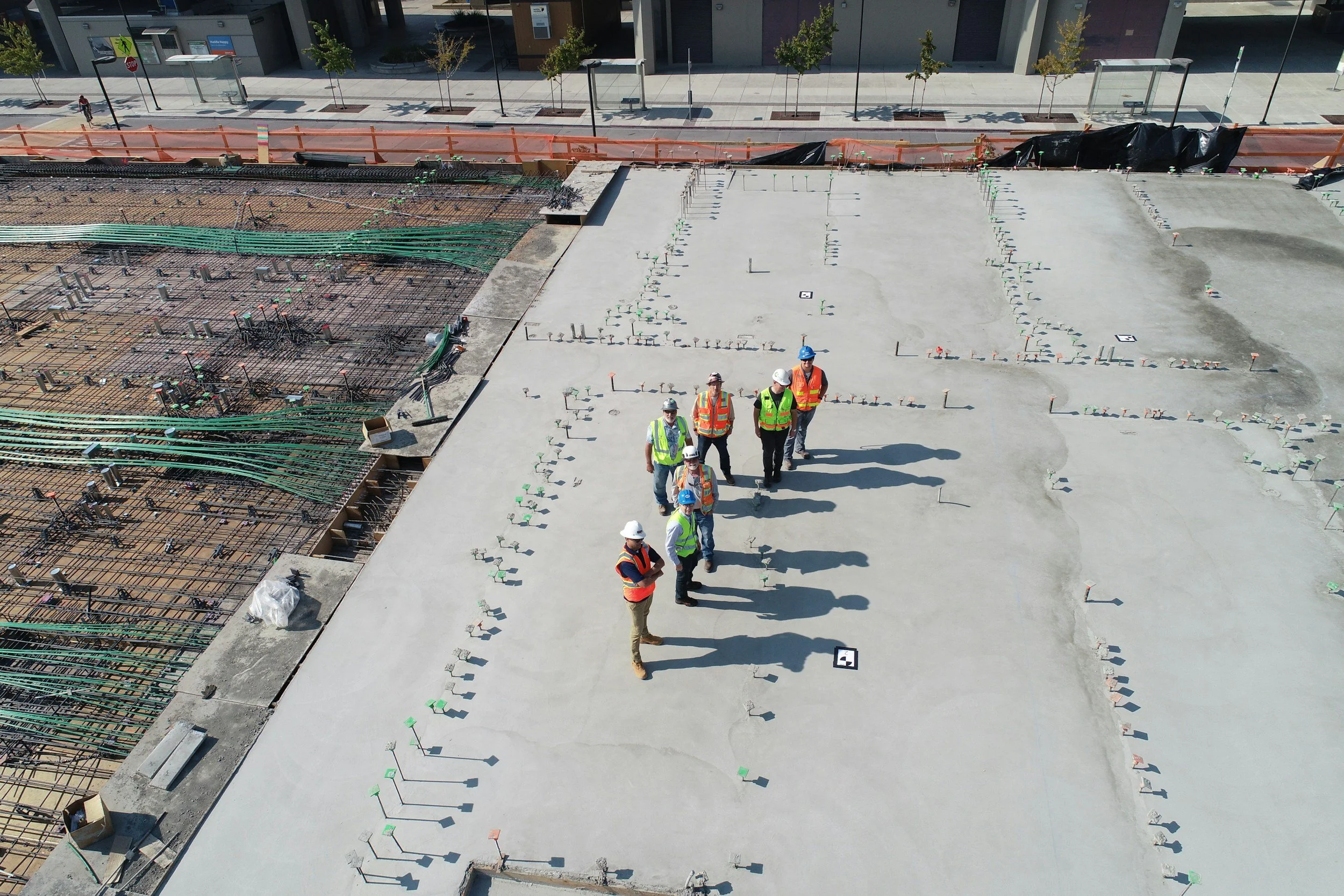 Group of construction workers wearing safety vests and helmets standing on freshly poured concrete at a construction site. The site has various construction materials and rebar visible, with a city street and buildings in the background.