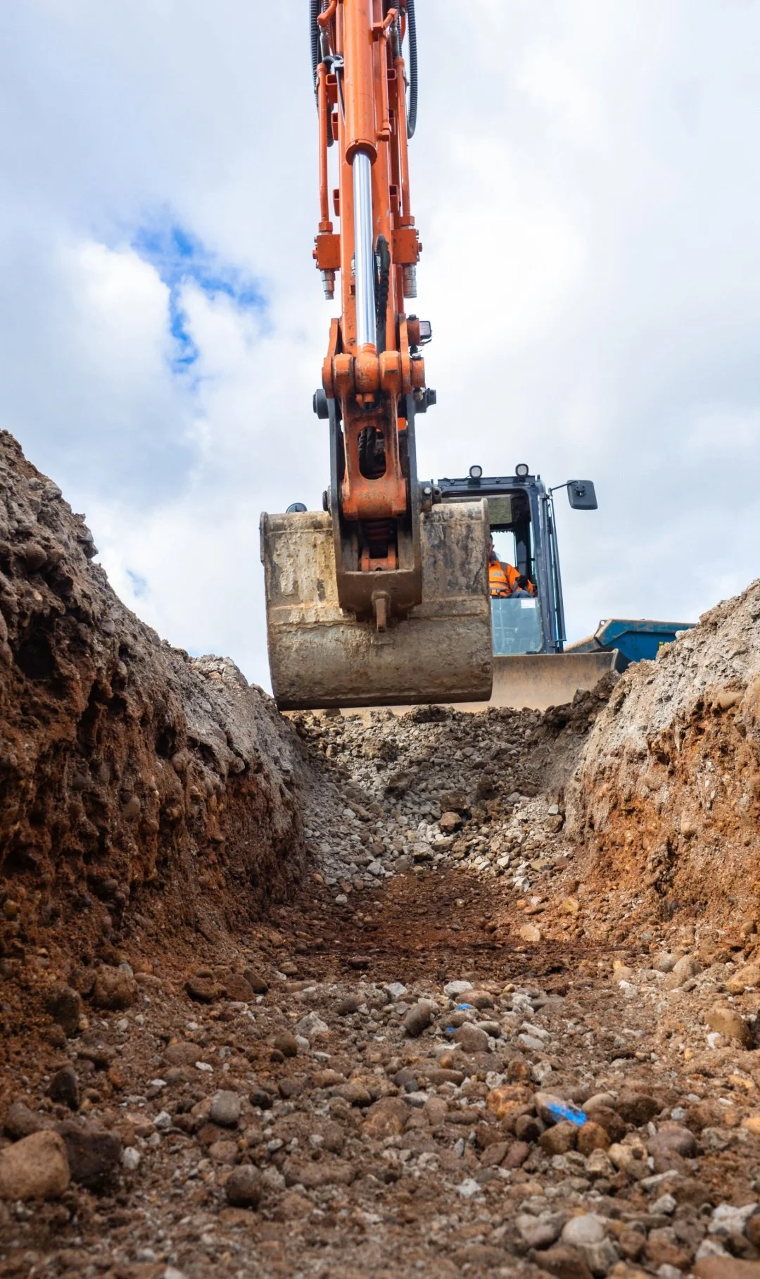 View from ground level showing a construction excavator digging in the dirt, with the sky and clouds in the background.