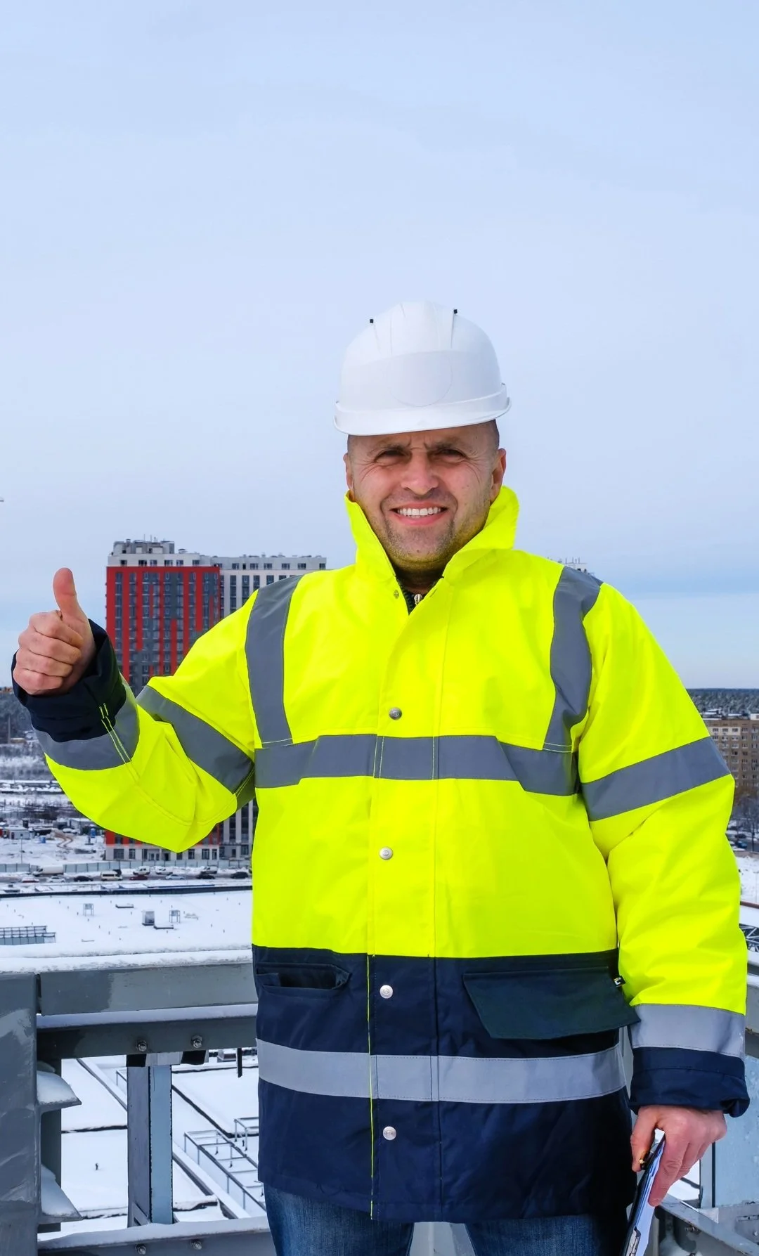 A construction worker wearing a yellow high-visibility jacket, a white hard hat, and holding a clipboard, standing outdoors on a snowy day with buildings in the background.