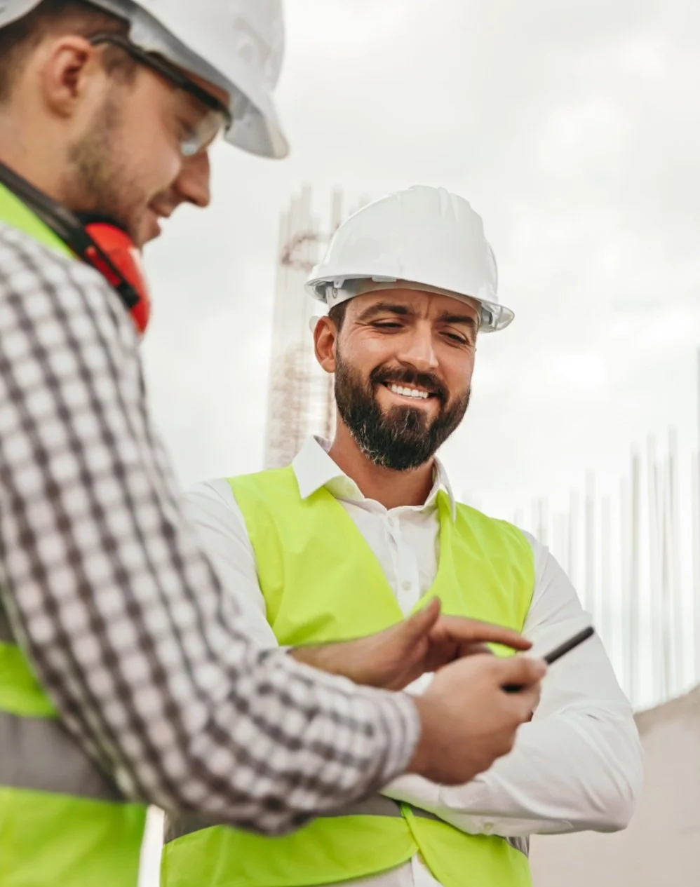 Two construction workers wearing hard hats and safety vests looking at a smartphone on a construction site.
