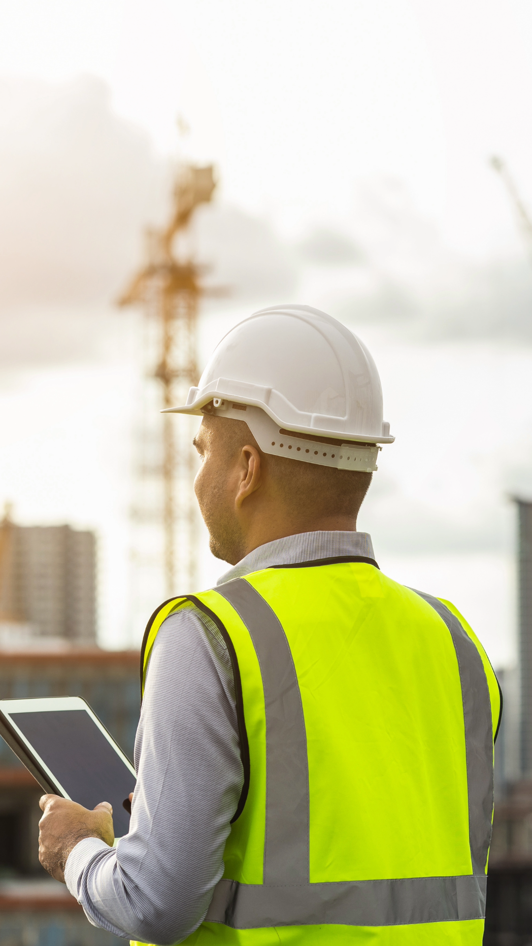A construction worker wearing a white safety helmet and a bright yellow reflective safety vest stands outside, holding a tablet and looking at a construction site with cranes and buildings in the background.
