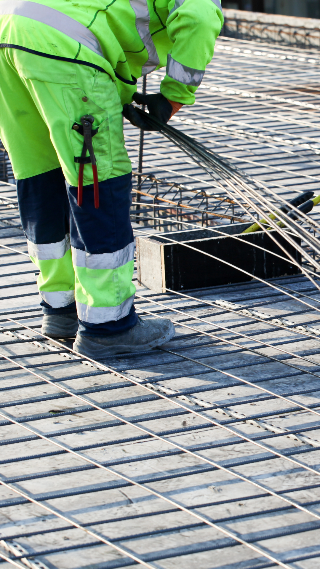 Construction worker in high-visibility safety gear working on a steel rebar framework at a construction site.