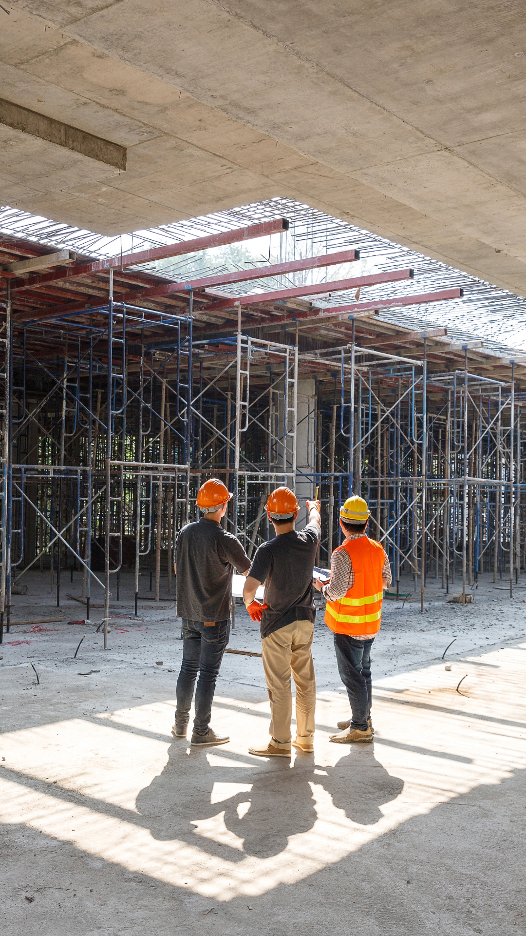 Three construction workers wearing helmets and safety vests discussing at a building site with scaffolding.