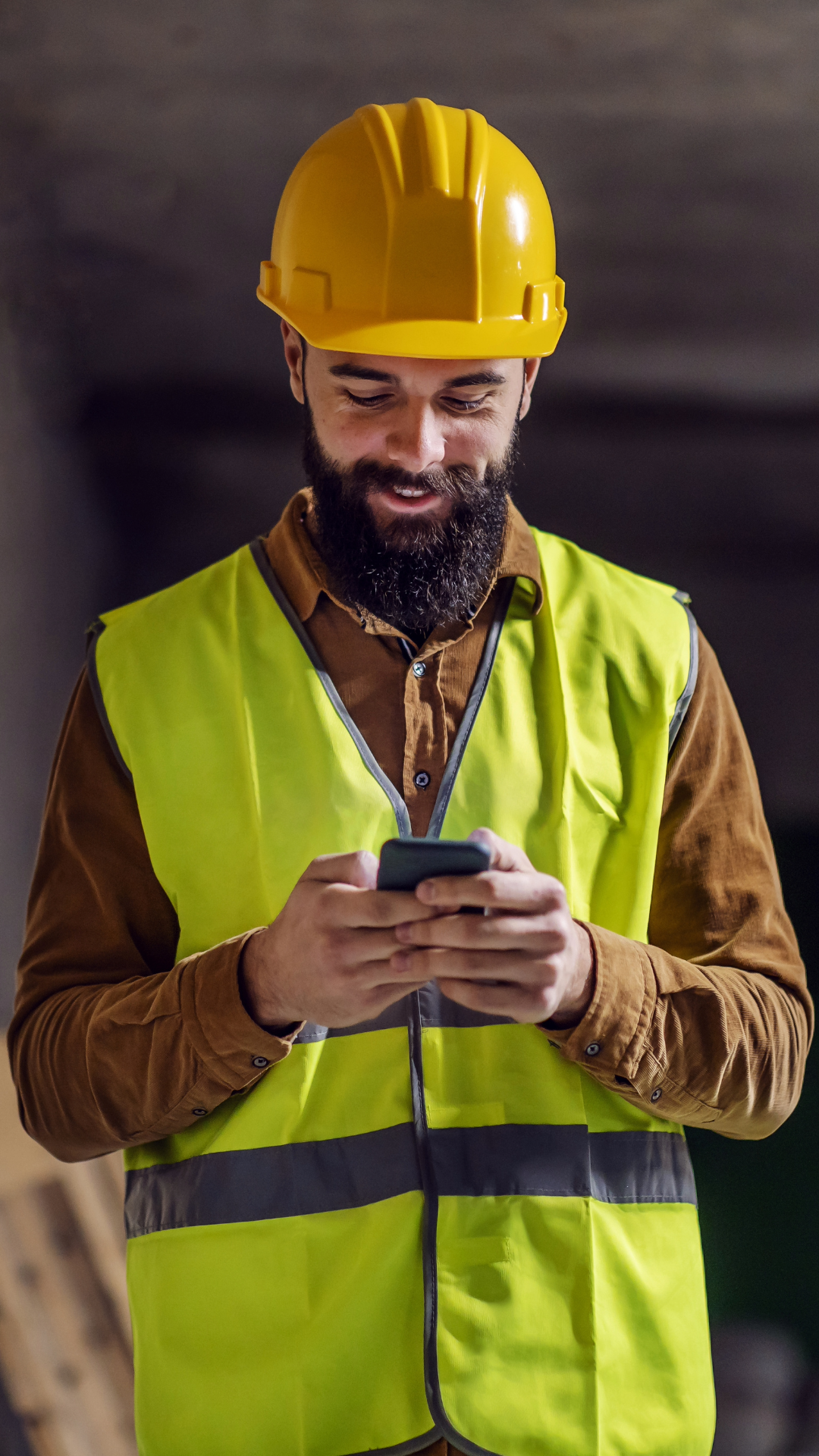 A construction worker wearing a yellow safety helmet and high-visibility vest, looking at a smartphone.