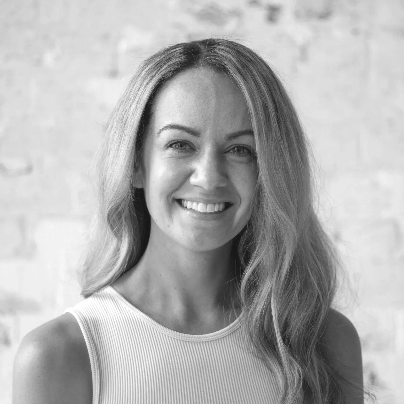 Black and white portrait of a smiling woman with long wavy hair, wearing a sleeveless top, standing against a brick wall background.