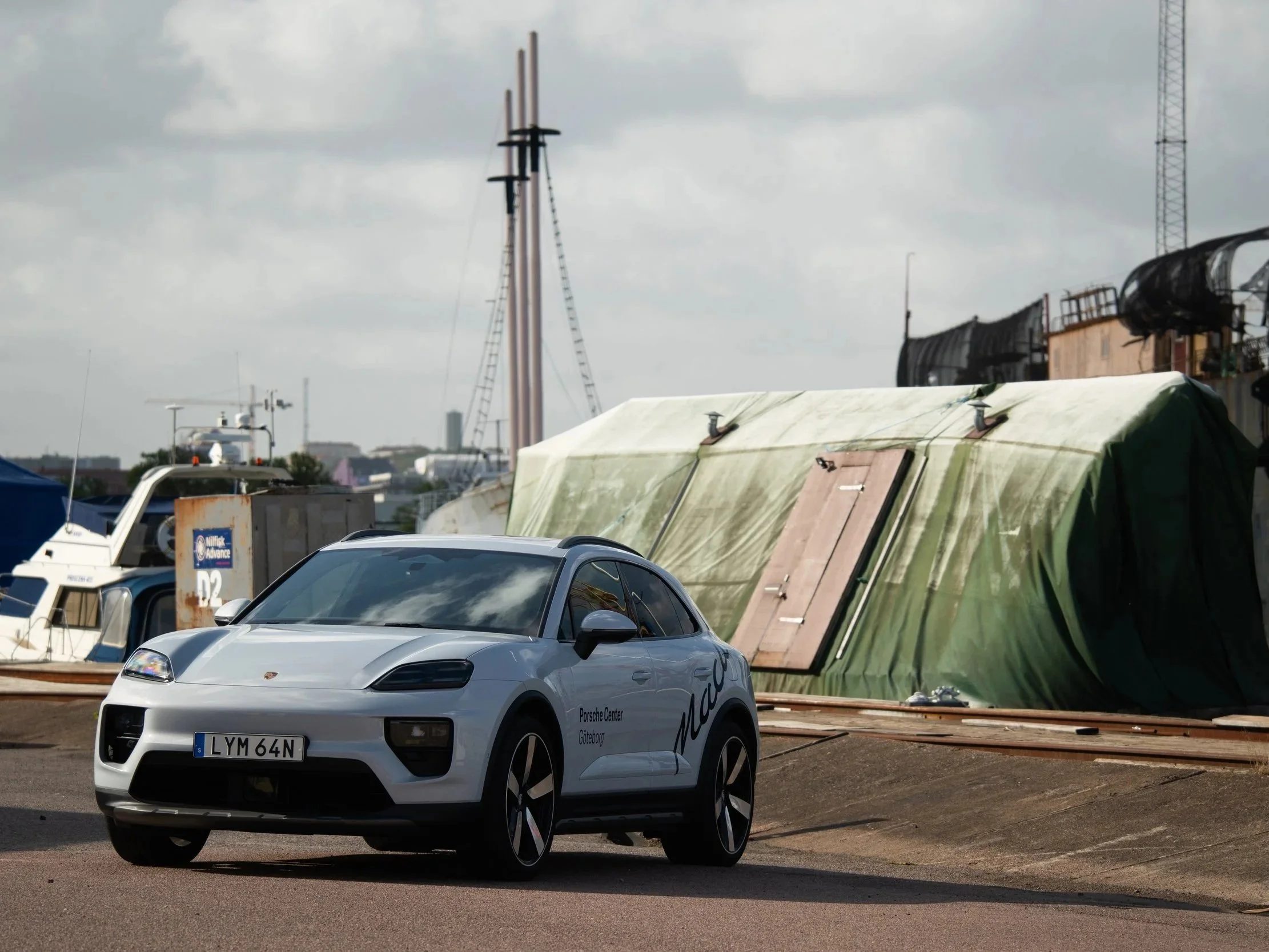 En vit Porsche Elcykelparkering står på en gata med båtar och en byggnad med grönt tält framför.