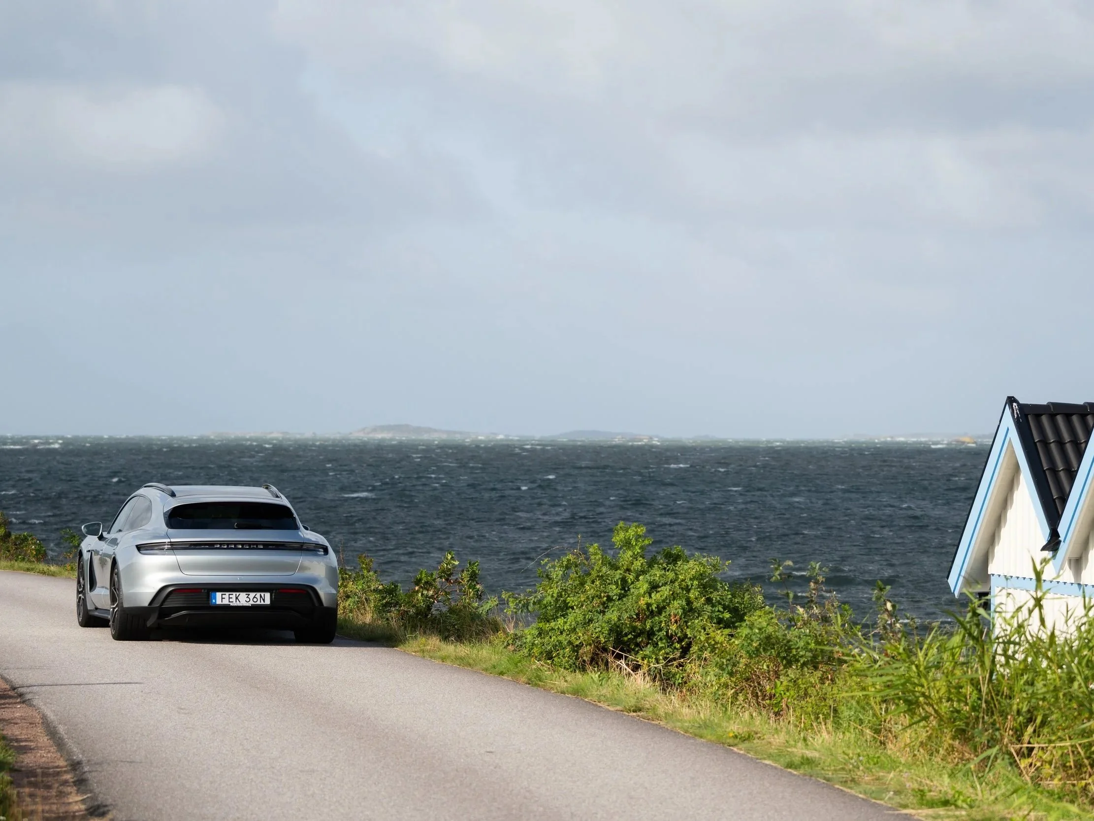 En silverfärgad sportbil parkerad bredvid en väg med havet i bakgrunden och ett grönskande område.