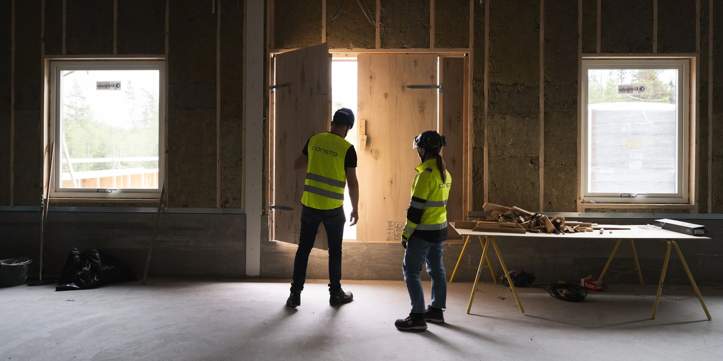 Två personer i byggnadsarbetsskydd, med hjälm och reflexväst, står inne i ett byggprojekt och diskuterar, med lägre hyllor av trä och byggmaterial på en arbetsbänk framför dem.