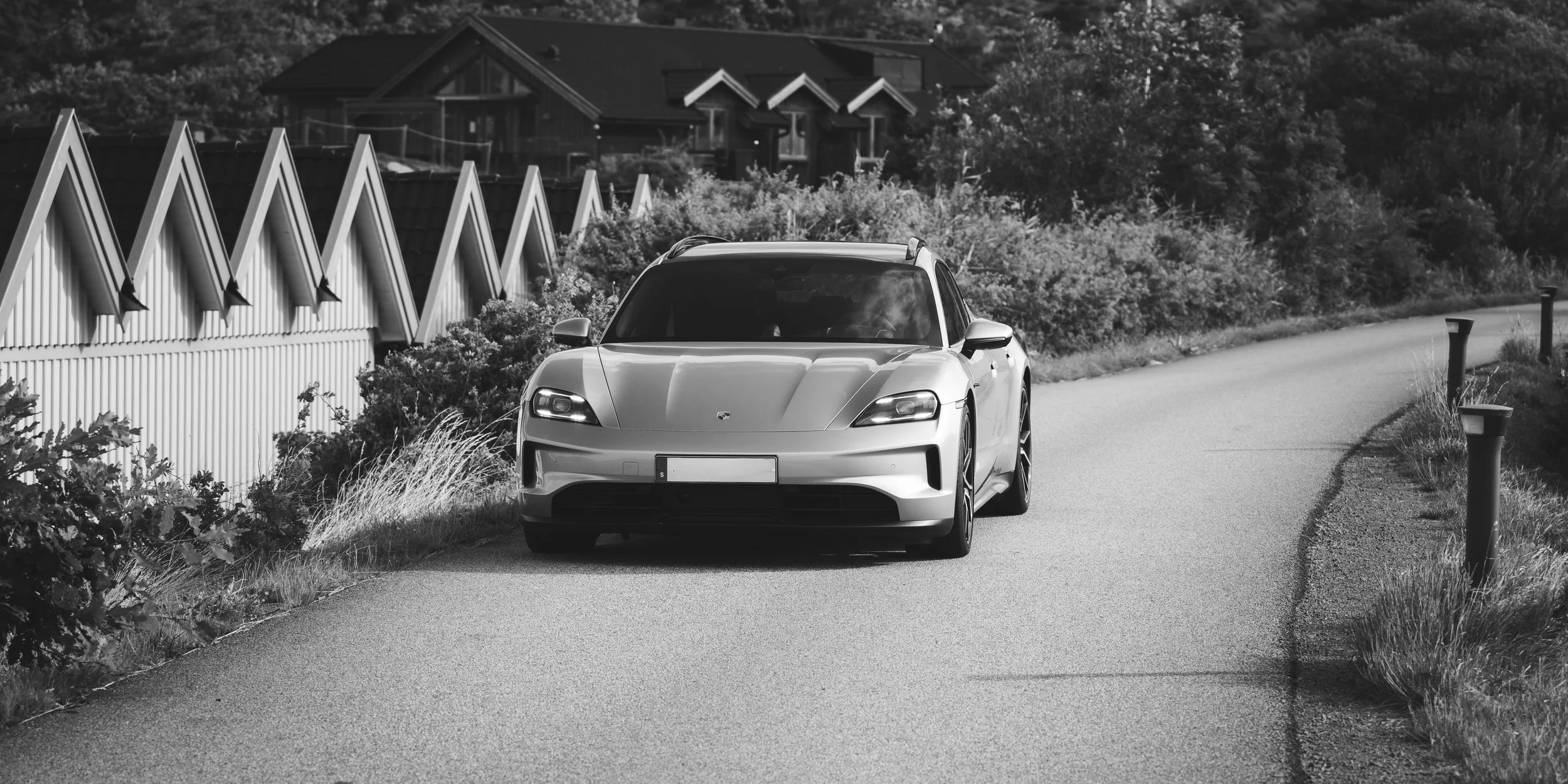 Svartvit bild av en modern bil parkerad längs en landsväg med hus och träd i bakgrunden.