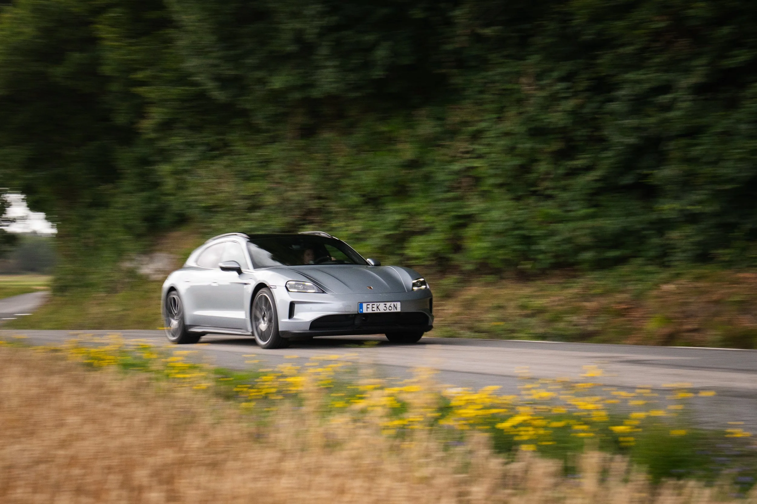 En silverfärgad Porsche kör på en landsväg med träd i bakgrunden och blommor i förgrunden.