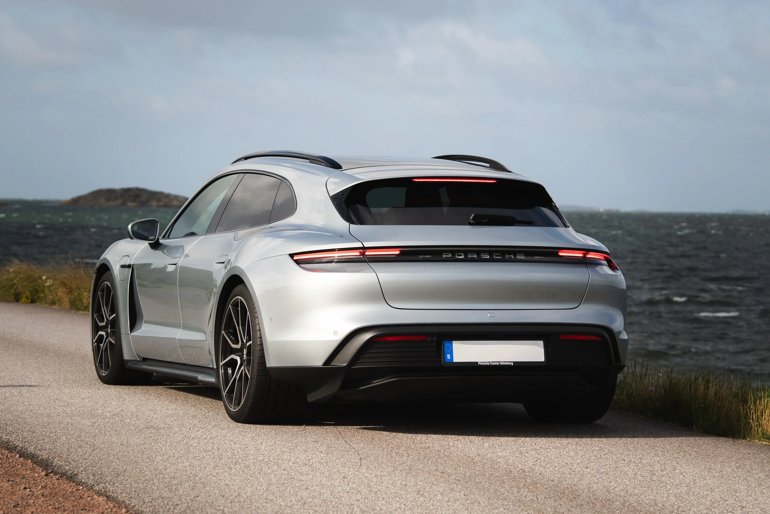 En silverfärgad Porsche-sedan är parkerad längs en kustväg med hav och öar i bakgrunden.