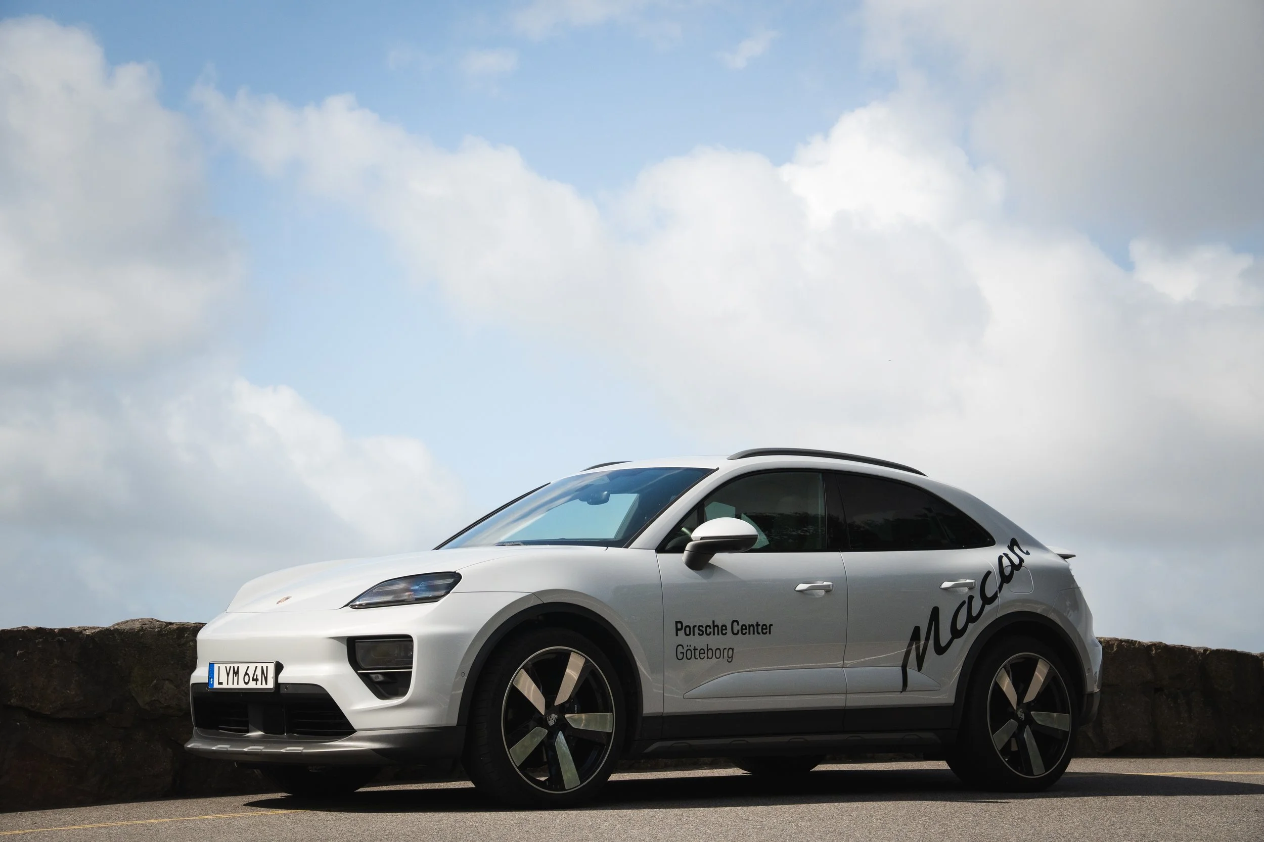 En vit Porsche Macan med texten 'Porsche Center Göteborg' på dörren, parkerad vid en stenmur under en molnig himmel.
