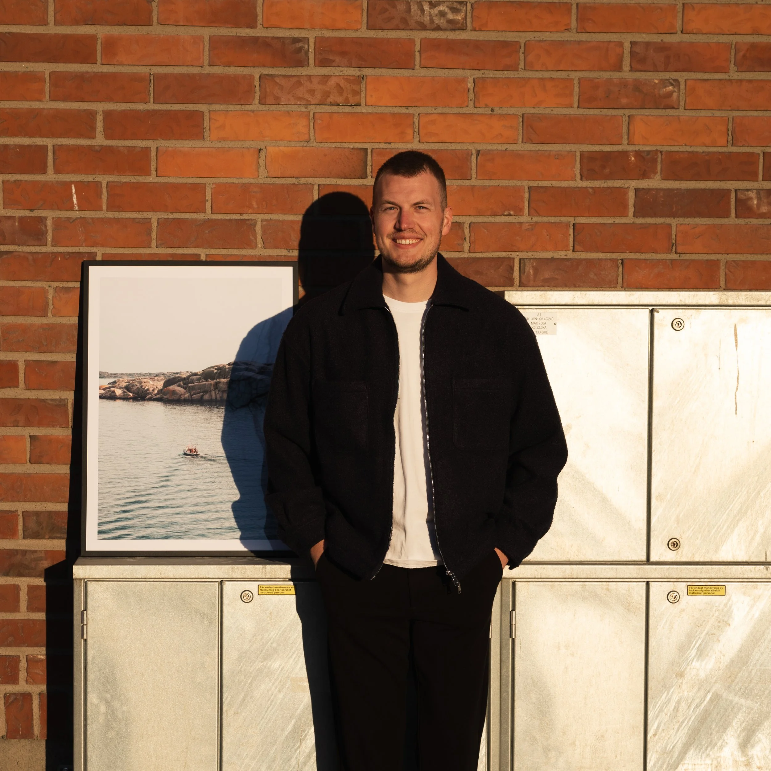 Porträtt av en leende man i svart jacka framför en tegelvägg, delvis skuggad av solljus.