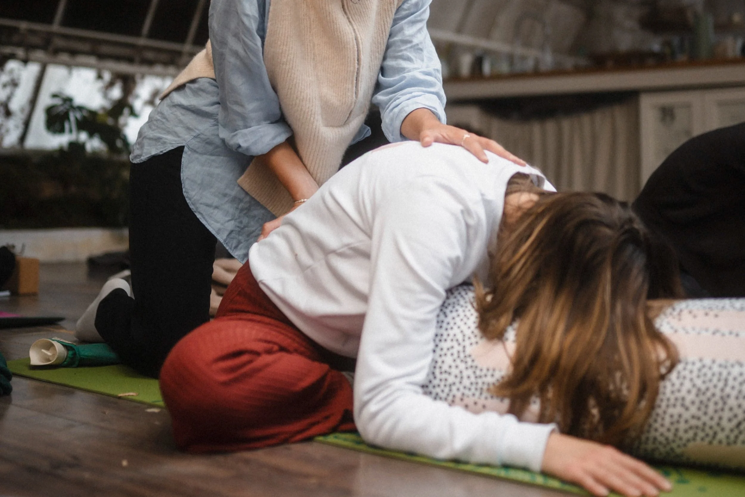 Eine Person in entspannter Yoga-Position auf einer Matte mit Unterstützung durch einen anderen, der auf dem Boden kniet.