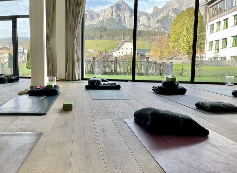 Innenansicht eines hellen Yoga-Studios mit Matten und Kissen auf dem Boden, große Fenster mit Blick auf eine idyllische Berglandschaft im Hintergrund. Holzboden und frische, grüne Umgebung sichtbar.
