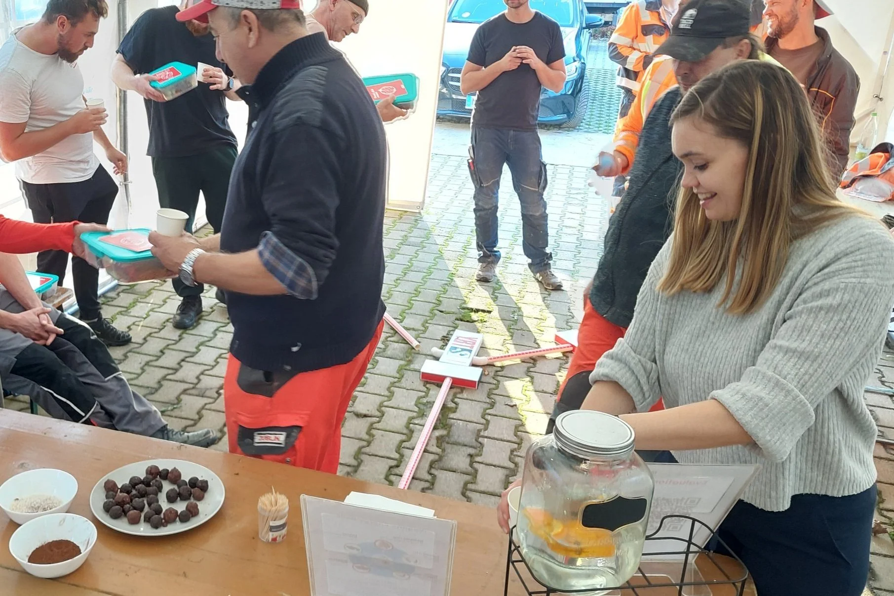 Menschen in einer Versammlung mit Essen und Getränken, mit Bauarbeitern in Arbeitskleidung und einer jungen Frau, die Getränke serviert. Im Vordergrund sind Schüsseln mit Snacks und ein Wasserspender zu sehen.