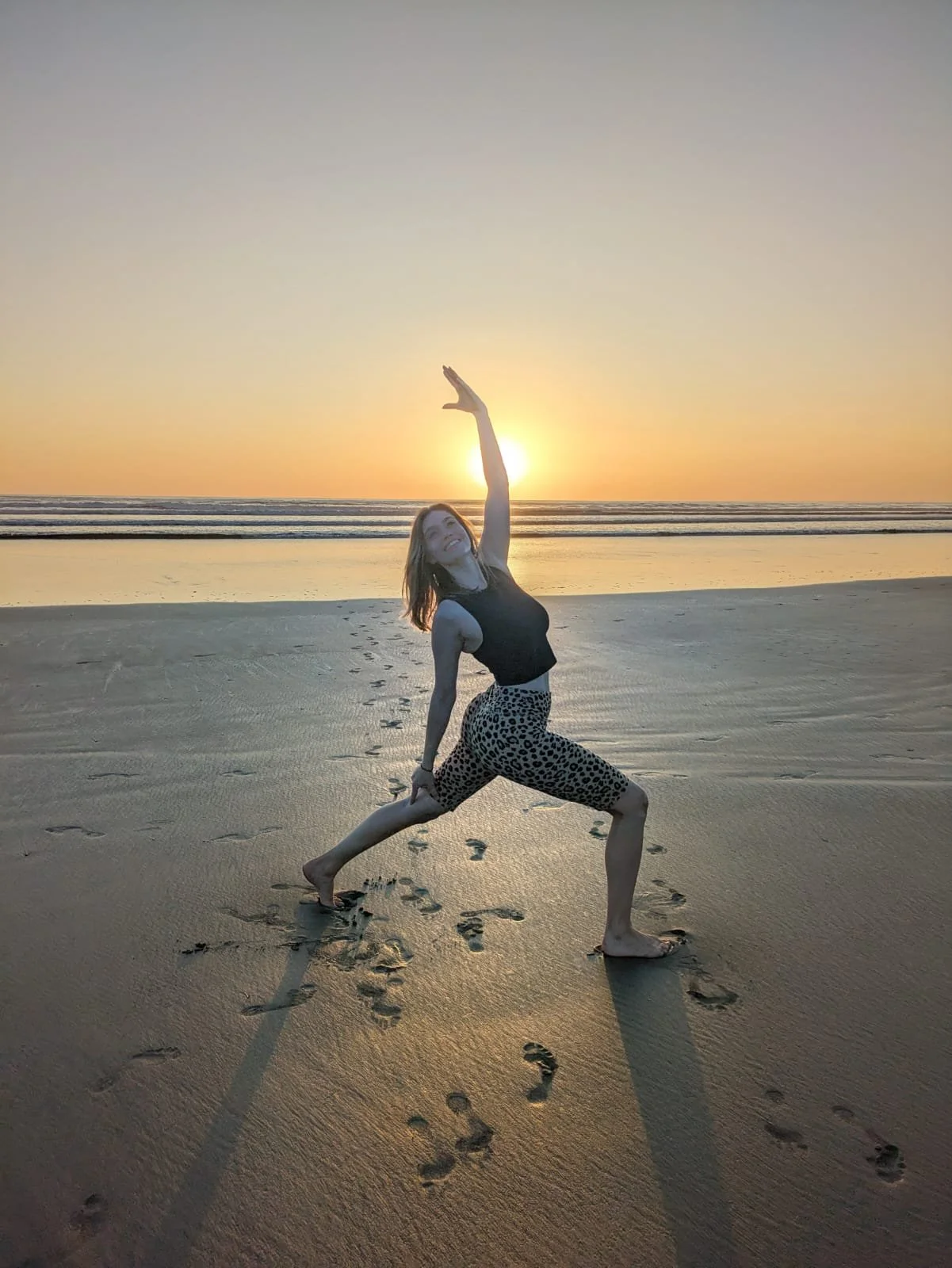 Frau macht Yoga am Strand bei Sonnenuntergang, Fußspuren im Sand sichtbar.