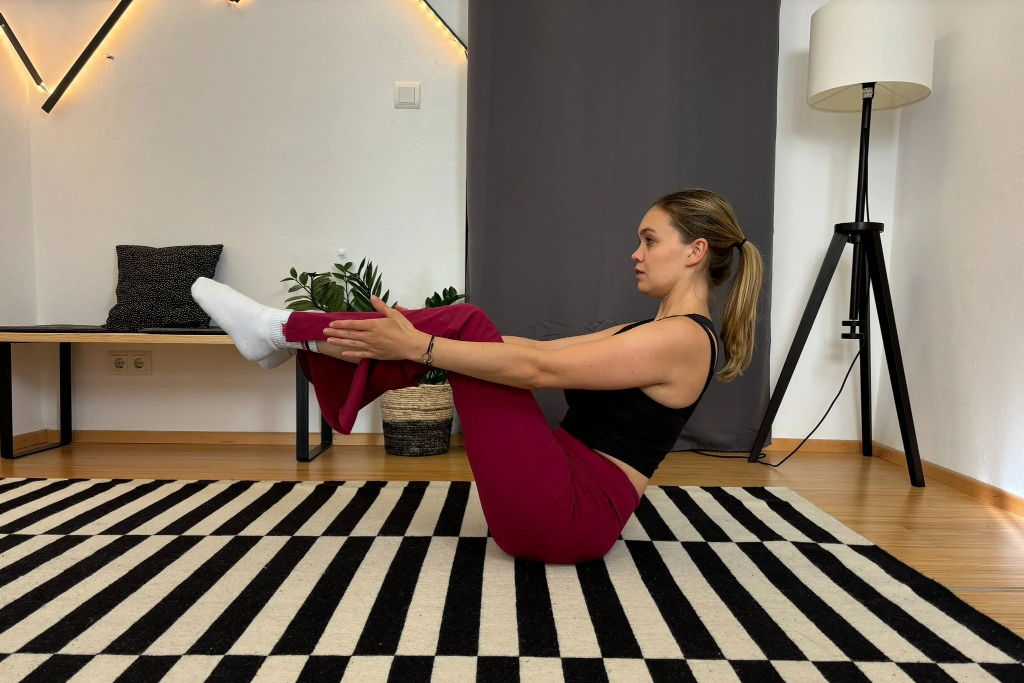 Frau macht Yoga in einem Raum, sie befindet sich in der Boot-Haltung auf einem gestreiften Teppich, im Hintergrund sind eine Pflanze, ein Sofa, und eine Stehlampe zu sehen.
