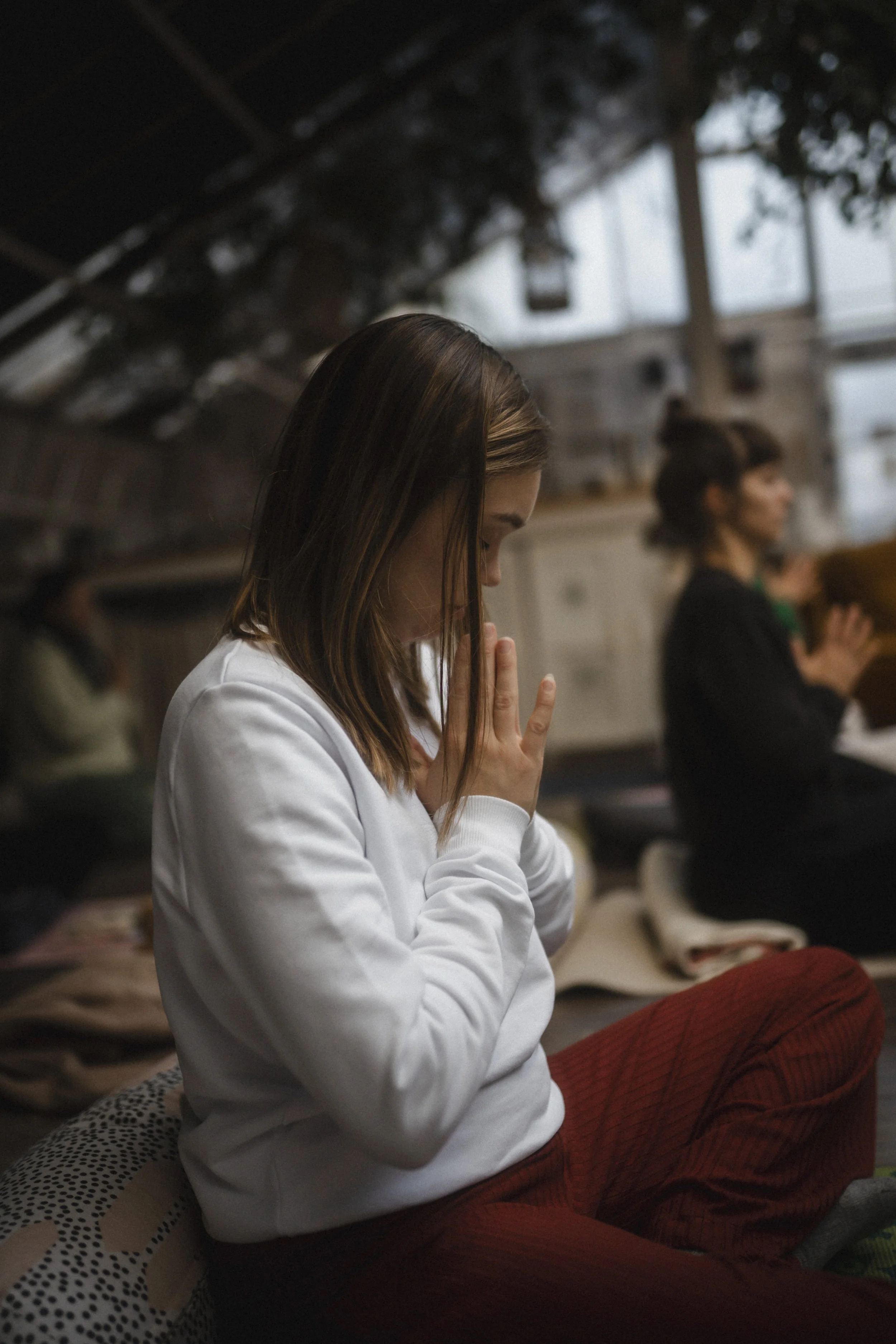 Eine Frau in einem weißen Pullover und roten Hosen sitzt in einem ruhigen Raum und hält ihre Hände in einer Gebetshaltung vor ihrem Gesicht. Sie scheint zu meditieren oder beten. Im Hintergrund sind weitere unscharfe Personen zu sehen, die in einer ähnlichen Haltung sind.