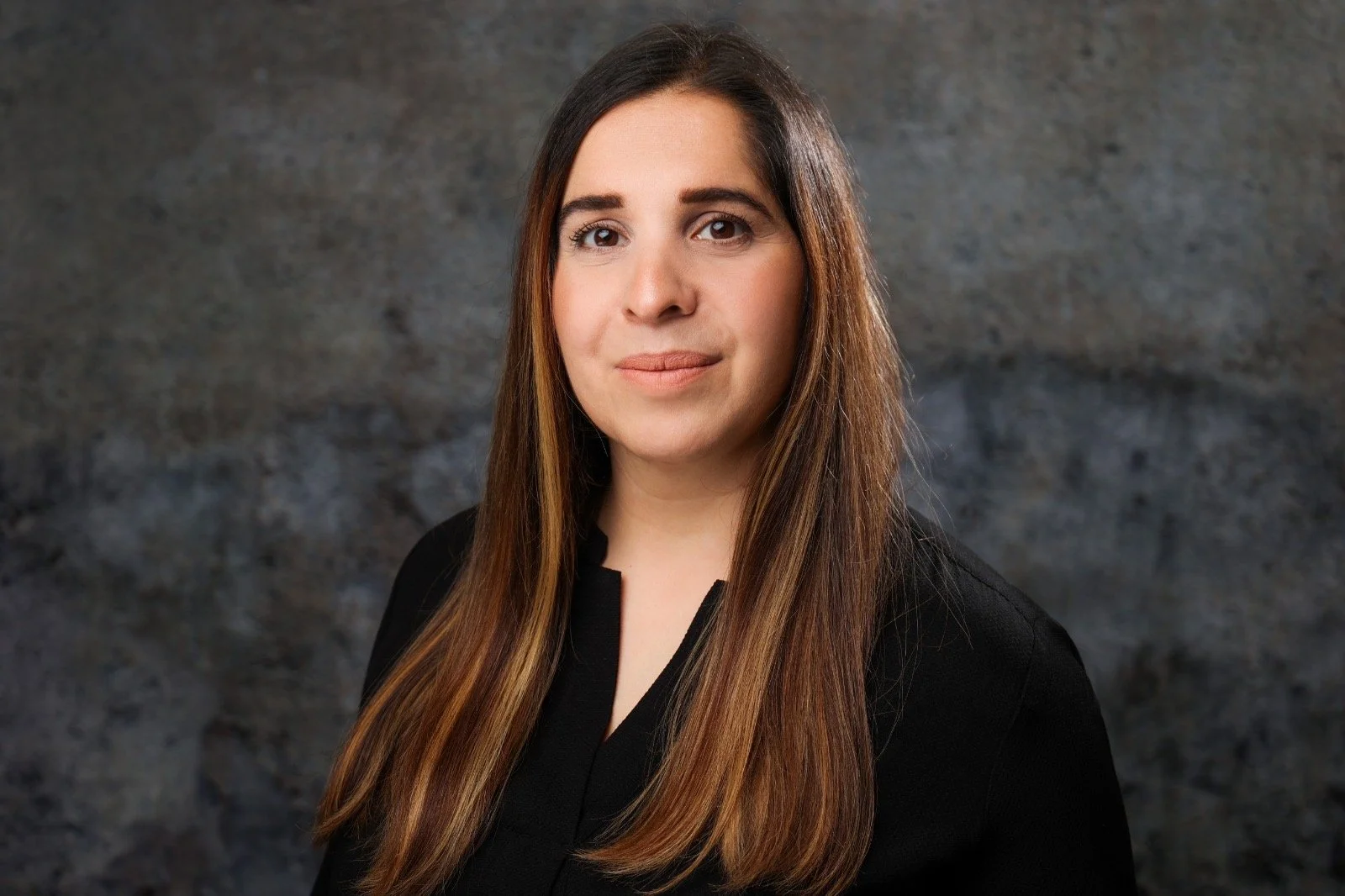 Porträt einer jungen Frau mit langen braunen Haaren, schwarzem Kleid, vor grauem Hintergrund.