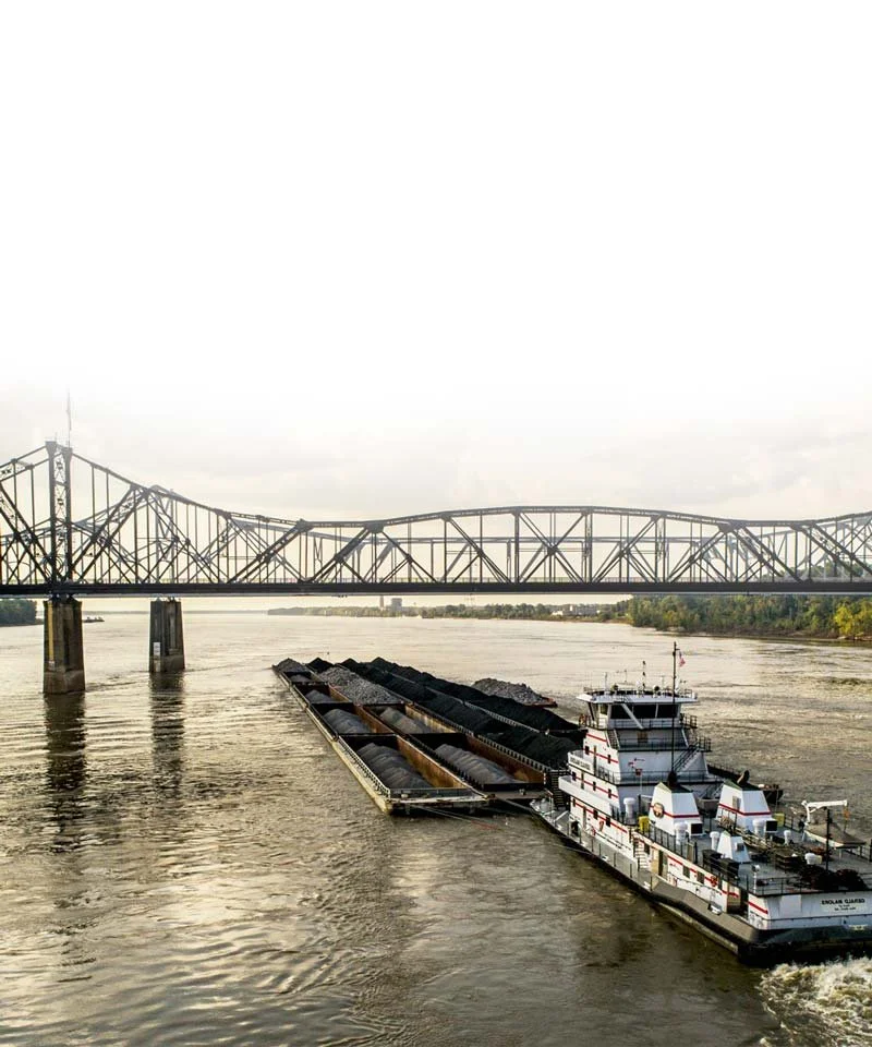 A barge carrying black coal moving on a river with a bridge in the background.