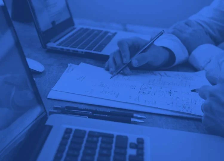 Two laptops, a notebook with handwritten notes, and two pens on a desk, with a person's hand writing in the notebook.